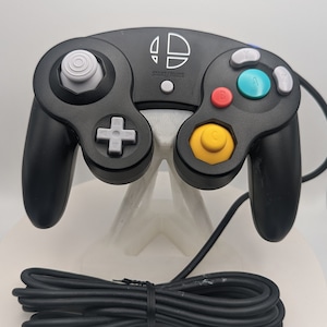 May include: A black wired video game controller featuring a white Super Smash Bros logo, a white analogue stick, a grey D-pad, a yellow C-stick, a red B button, a green A button, and grey X, Y, and Z buttons, with a long black cable coiled at the base.
