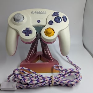 Op de afbeelding: Een witte Nintendo GameCube-controller met paarse knoppen, een gele C-stick en een grijze analoge stick. De controller staat op een roze en oranje standaard en heeft een gevlochten witte, paarse en roze kabel met een witte connector.