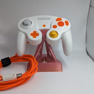 May include: A white Nintendo GameCube controller featuring orange action buttons, a yellow C-stick, a gray analog stick, and a gray D-pad. The controller is mounted on a pink stand and includes a long, coiled orange cable with a white connector plug.