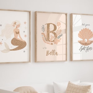 Peut inclure: Trois impressions encadrées sur le thème de la plage. La première impression représente une sirène aux longs cheveux blonds et à la queue brune. La deuxième impression représente la lettre B avec un motif floral et le texte "is for Bella". La troisième impression représente un coquillage avec le texte "Let your light shine".