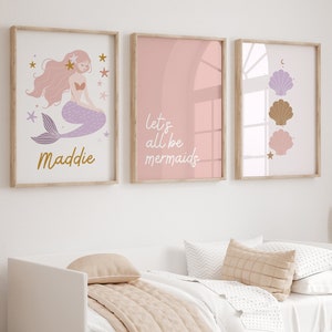 Puede incluir: Tres impresiones enmarcadas con fondo rosa. La impresión de la izquierda presenta una sirena morada con cabello rosado y estrellas doradas. La impresión del medio dice "Let's all be mermaids". La impresión de la derecha presenta tres conchas en tonos de morado, marrón y rosa.