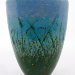 May include: A blue and green ceramic vase with a painted design of a field of wildflowers. The vase has a wide mouth and a narrow base.