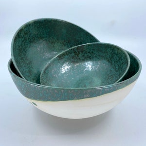 May include: Three ceramic bowls in various sizes, nested together. The bowls are a deep teal green with a white rim and a speckled glaze.