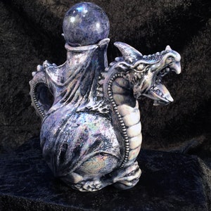Può includere: Una statuetta di drago in ceramica con smalto blu e nero iridescente. Il drago ha una grande bocca aperta e una sfera di vetro viola sulla testa.