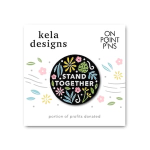 Könnte beinhalten: Schwarze Emaille-Anstecknadel mit floralem Design und dem Text "STAND TOGETHER". Die Anstecknadel ist von einem weißen Rand umgeben und hat einen schwarzen Hintergrund. Die Anstecknadel ist Teil einer Kollektion von Anstecknadeln von kela designs.