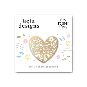 Puede incluir: Pin de esmalte dorado en forma de corazón con diseños florales blancos y el texto "Proudly Asian". El pin está sobre un fondo blanco con un diseño floral y el texto "kela designs" y "ON POINT PINS".