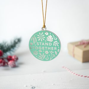 May include: A round, mint green ornament with the words "STAND TOGETHER" in white, surrounded by floral and leaf designs. The ornament hangs from a gold string. A gift box and festive greenery are in the background.