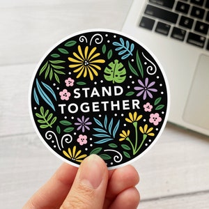 Puede incluir: Una pegatina redonda en blanco y negro con un diseño floral y el texto "STAND TOGETHER". La pegatina tiene un borde blanco y está siendo sostenida por una mano.