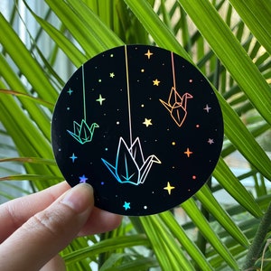 Puede incluir: Una pegatina redonda con fondo negro y grullas de origami de arcoíris iridiscente colgando de cuerdas sobre un fondo de estrellas.
