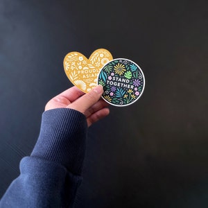 K&ouml;nnte beinhalten: Zwei Aufkleber mit Blumenmustern. Ein Aufkleber ist herzf&ouml;rmig mit dem Text "Proudly Asian" und der andere ist ein Kreis mit dem Text "Stand Together".