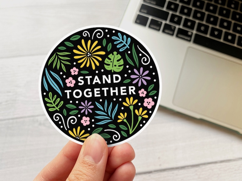 K&ouml;nnte beinhalten: Ein runder Aufkleber in Schwarzwei&szlig; mit einem floralen Design und dem Text "STAND TOGETHER". Der Aufkleber hat einen wei&szlig;en Rand und wird von einer Hand gehalten.