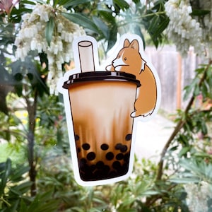 Op de afbeelding: Een sticker van een bruine en witte corgi-hond die een bruine en witte boba-theebeker vasthoudt met een zwarte rietje. De boba-theebeker is gevuld met bruine thee en zwarte boba-parels.