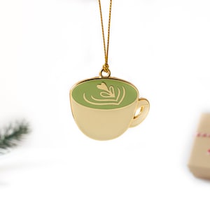 May include: Gold-colored ornament shaped like a cup of green matcha latte with a leaf design. The ornament is hanging from a gold string.