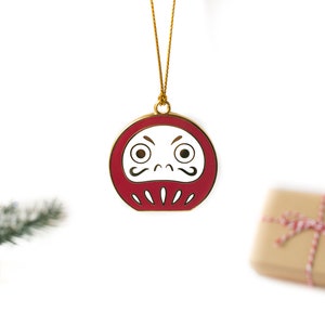 May include: A red and gold Christmas ornament shaped like a Japanese Daruma doll with a white face and black eyes and mustache. The ornament is hanging from a gold string.