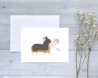 May include: A white greeting card with a cartoon corgi illustration. The corgi, in black, brown, and white, carries flowers. Text below reads "FOR YOU~". A white envelope is on the left. The background is a light grey wooden surface.