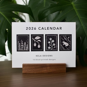 May include: A 2026 calendar with four black and white block-printed designs, including botanical and animal illustrations. The calendar is displayed on a wooden stand, with the text "2026 Calendar" and "Kela Designs".
