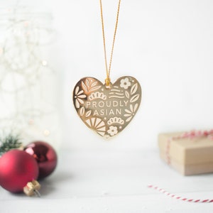 May include: Gold heart-shaped ornament with a white floral design and the text "Proudly Asian".