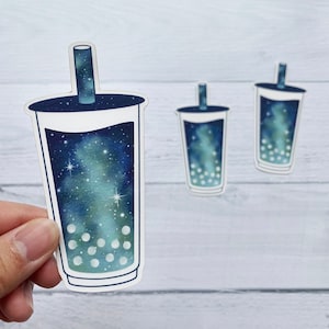 May include: Three galaxy-themed bubble tea stickers with a blue and green galaxy design and white boba pearls. The stickers are on a white wooden background.