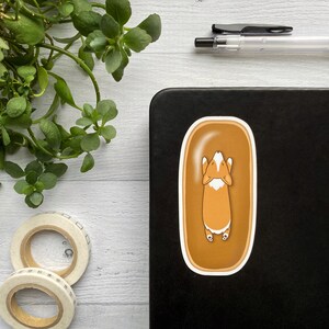 May include: A sticker of a corgi dog laying on its back in a brown bathtub. The corgi is orange and white with its paws in the air.