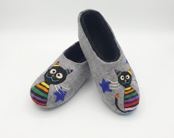 May include: A pair of gray felt slippers with a black cat design. The cat has a rainbow striped body, a colorful hat, and is surrounded by blue stars. The slippers have a black interior.