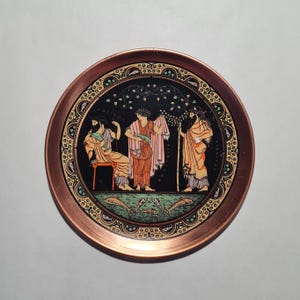 May include: A round copper-toned metal tray featuring a central black circular illustration of three figures in classical robes under a tree, surrounded by a decorative border with small fish motifs and the text Poseidon, Amphitrite, and Dioniso.