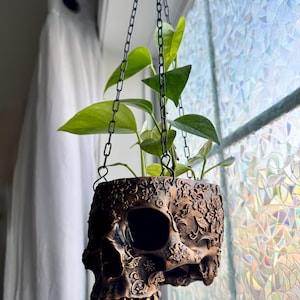 May include: A hanging planter shaped like a human skull with a dark bronze finish and intricate floral relief carvings. The skull features deep eye sockets and a nasal cavity, with a green leafy plant growing from the top, suspended by black chains.