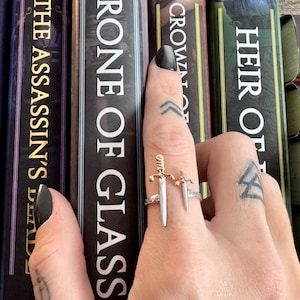 Puede incluir: Un anillo plateado con dos colgantes de espada, uno más grande que el otro, se muestra frente a una pila de libros. El anillo está en un dedo con esmalte de uñas negro y un tatuaje. Los títulos de los libros incluyen "Throne of Glass."