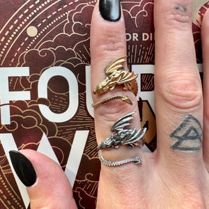 May include: Three dragon-themed rings in gold and silver tones. The rings are worn on a hand with black nail polish. The rings feature detailed dragon designs, with one ring having a tail of small stones.