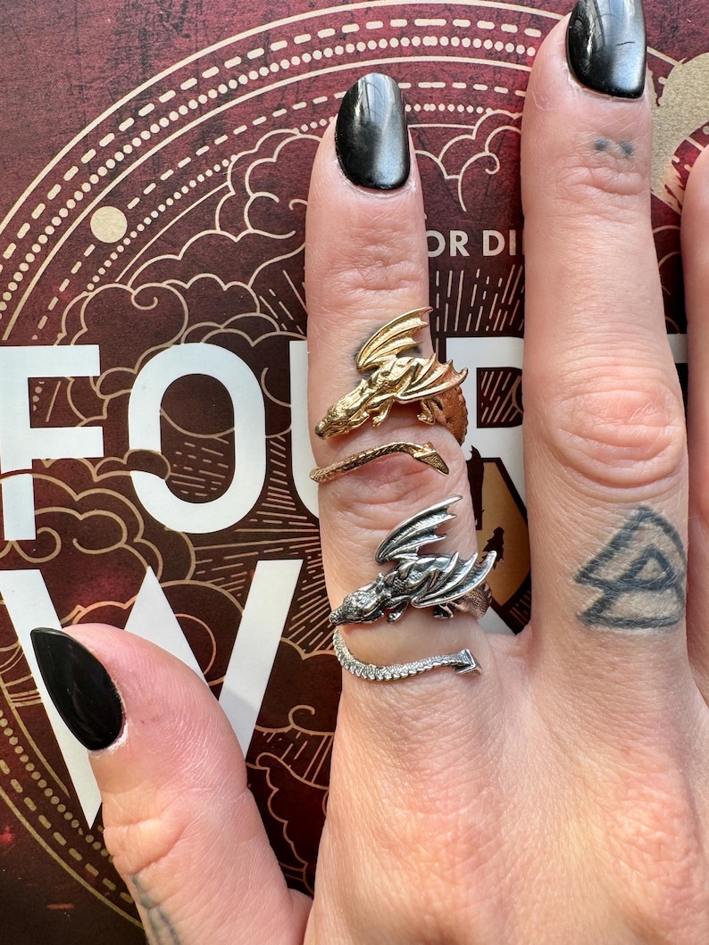 May include: Three dragon-themed rings in gold and silver tones. The rings are worn on a hand with black nail polish. The rings feature detailed dragon designs, with one ring having a tail of small stones.