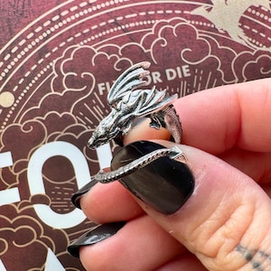 May include: A silver dragon ring with intricate detailing. The dragon's head and wings are prominently displayed, wrapping around the finger. The ring is worn on a hand with black nail polish. The background has a decorative design with the words "Fly or Die".