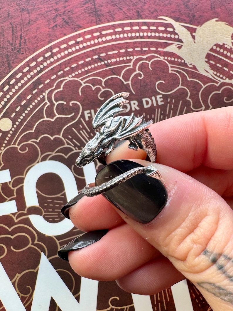 May include: A silver dragon ring with intricate detailing. The dragon's head and wings are prominently displayed, wrapping around the finger. The ring is worn on a hand with black nail polish. The background has a decorative design with the words "Fly or Die".