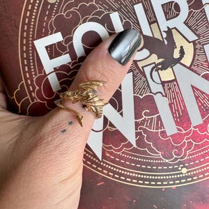 May include: A gold-colored dragon ring with intricate details, wrapped around a finger. The ring is set against a book cover with the words "FOURTH WING" and "FLY...OR DIE" in white. The background is a deep red with gold accents.