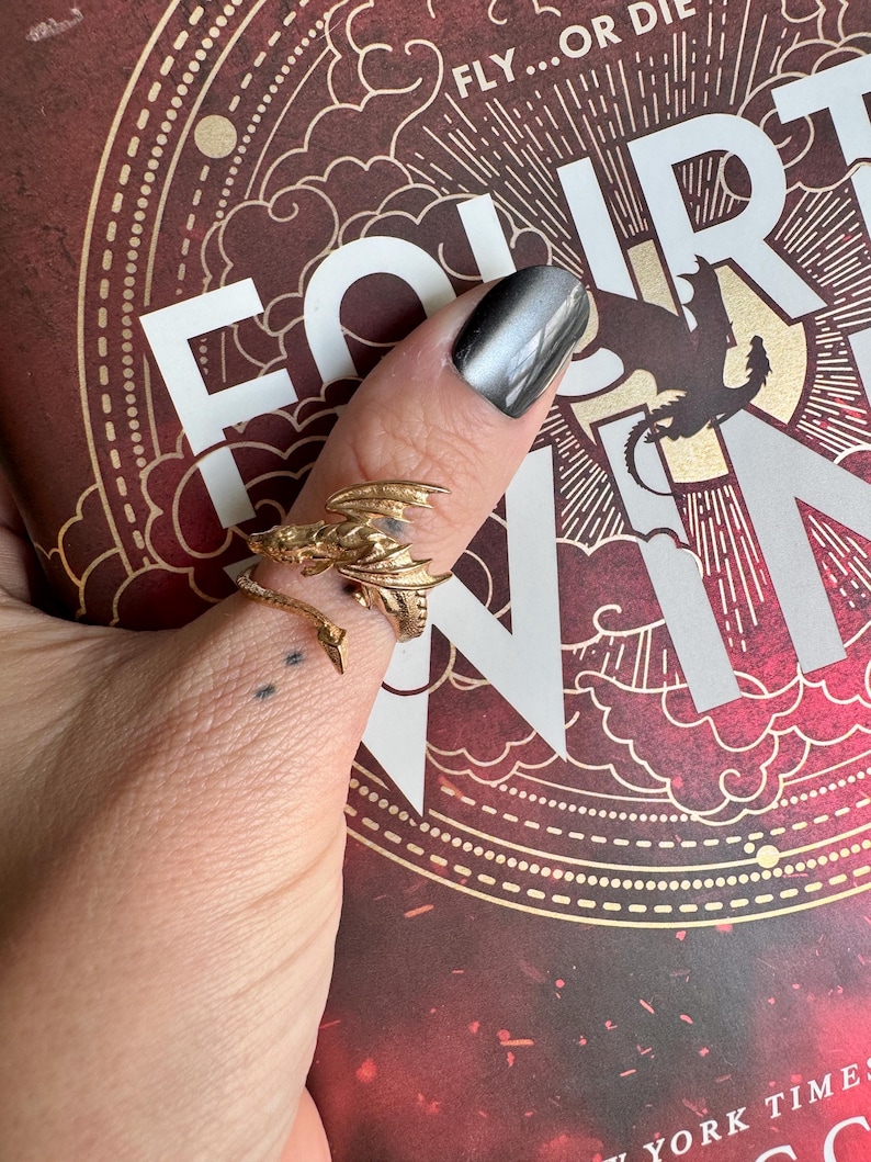 May include: A gold-colored dragon ring with intricate details, wrapped around a finger. The ring is set against a book cover with the words "FOURTH WING" and "FLY...OR DIE" in white. The background is a deep red with gold accents.