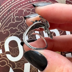 May include: A silver dragon ring with intricate detailing. The dragon's head and tail curve to form the ring's design. The ring is held between fingers with black nail polish, showcasing the ring's design.