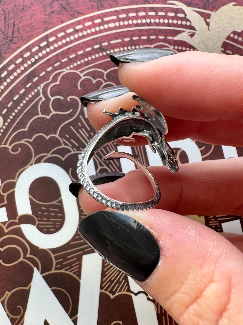 May include: A silver dragon ring with intricate detailing. The dragon's head and tail curve to form the ring's design. The ring is held between fingers with black nail polish, showcasing the ring's design.