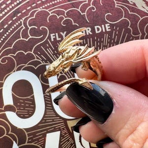 May include: A gold-colored dragon ring with detailed wings and scales. The ring wraps around the finger, with the dragon's head and tail meeting. The background features the words "FLY OR DIE" in a decorative font.
