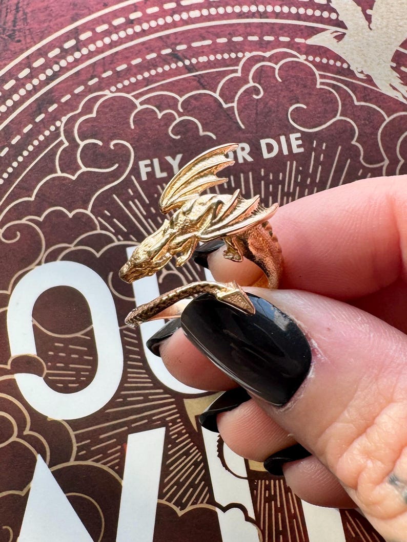 May include: A gold-colored dragon ring with detailed wings and scales. The ring wraps around the finger, with the dragon's head and tail meeting. The background features the words "FLY OR DIE" in a decorative font.