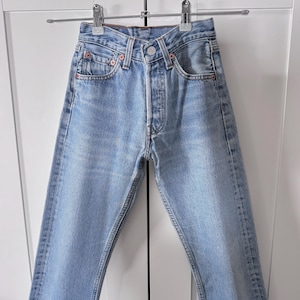 May include: Light blue denim jeans hanging on a metal hanger. The jeans have a button fly, classic five-pocket design, and red stitching details. The jeans are a light wash and appear to be new.