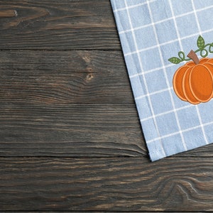 K&ouml;nnte beinhalten: Ein blaues und wei&szlig;es kariertes K&uuml;chentuch mit einer gestickten orangefarbenen K&uuml;rbis mit gr&uuml;nen Bl&auml;ttern.