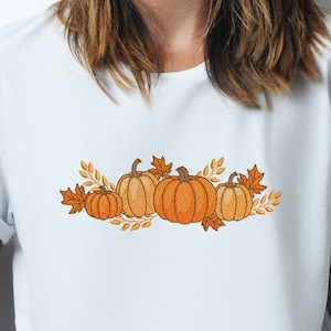 Op de afbeelding: Een wit T-shirt met een geborduurd ontwerp van oranje pompoenen en herfstbladeren.
