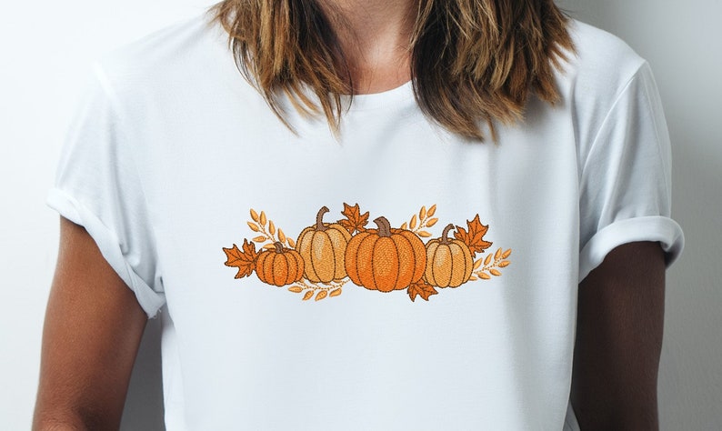 Op de afbeelding: Een wit T-shirt met een geborduurd ontwerp van oranje pompoenen en herfstbladeren.