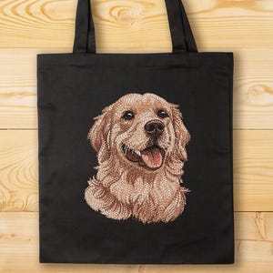 May include: Black tote bag with an embroidered Golden Retriever dog portrait. The dog has light brown fur and a pink tongue, and is smiling. The bag has black handles and is set against a wooden background.