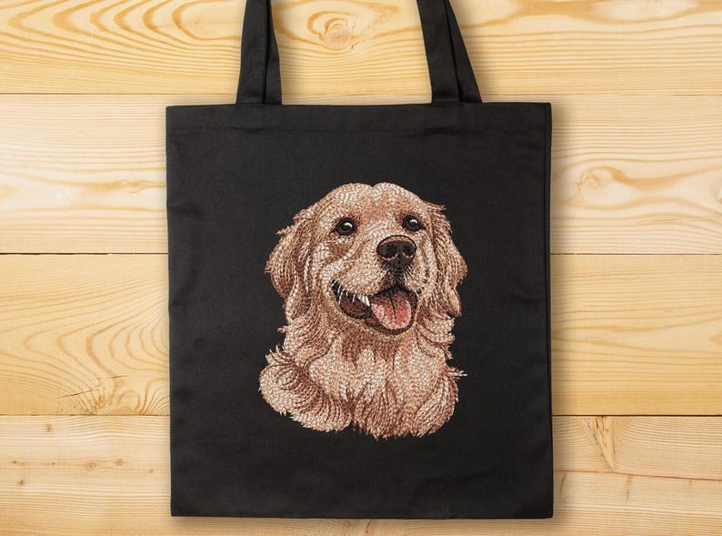 May include: Black tote bag with an embroidered Golden Retriever dog portrait. The dog has light brown fur and a pink tongue, and is smiling. The bag has black handles and is set against a wooden background.