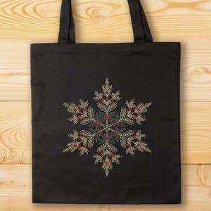 Puede incluir: Bolso tote negro con un dise&ntilde;o de copo de nieve bordado. El copo de nieve est&aacute; compuesto por elementos verdes, dorados y rojos, creando un tema invernal festivo. El bolso tiene dos asas negras y est&aacute; sobre un fondo de madera.