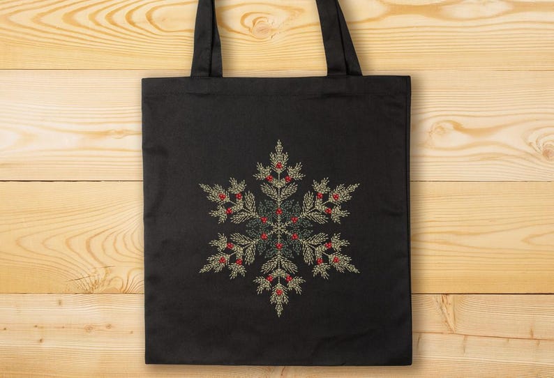 Puede incluir: Bolso tote negro con un dise&ntilde;o de copo de nieve bordado. El copo de nieve est&aacute; compuesto por elementos verdes, dorados y rojos, creando un tema invernal festivo. El bolso tiene dos asas negras y est&aacute; sobre un fondo de madera.