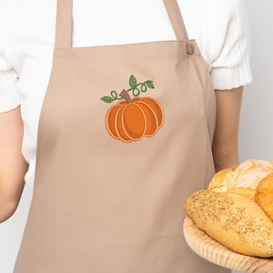 K&ouml;nnte beinhalten: Eine braune Sch&uuml;rze mit einer gestickten orangefarbenen K&uuml;rbis mit gr&uuml;nen Bl&auml;ttern. Eine Person h&auml;lt ein Holztablett mit verschiedenen Broten.
