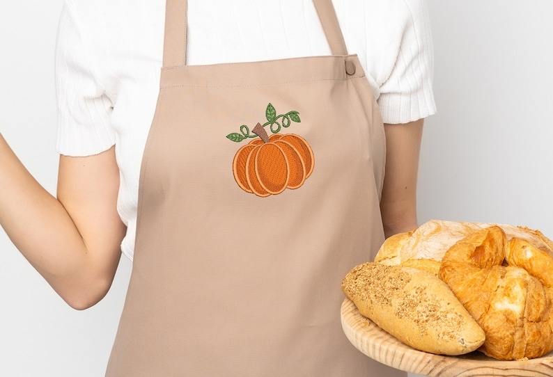 K&ouml;nnte beinhalten: Eine braune Sch&uuml;rze mit einer gestickten orangefarbenen K&uuml;rbis mit gr&uuml;nen Bl&auml;ttern. Eine Person h&auml;lt ein Holztablett mit verschiedenen Broten.