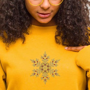 Puede incluir: Sudadera amarilla mostaza con un dise&ntilde;o detallado de copo de nieve y detalles de bayas rojas. El copo de nieve est&aacute; centrado en el pecho. La modelo lleva gafas con montura dorada. La sudadera tiene mangas largas y cuello redondo.