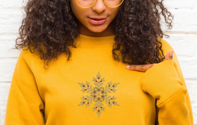 Puede incluir: Sudadera amarilla mostaza con un dise&ntilde;o detallado de copo de nieve y detalles de bayas rojas. El copo de nieve est&aacute; centrado en el pecho. La modelo lleva gafas con montura dorada. La sudadera tiene mangas largas y cuello redondo.