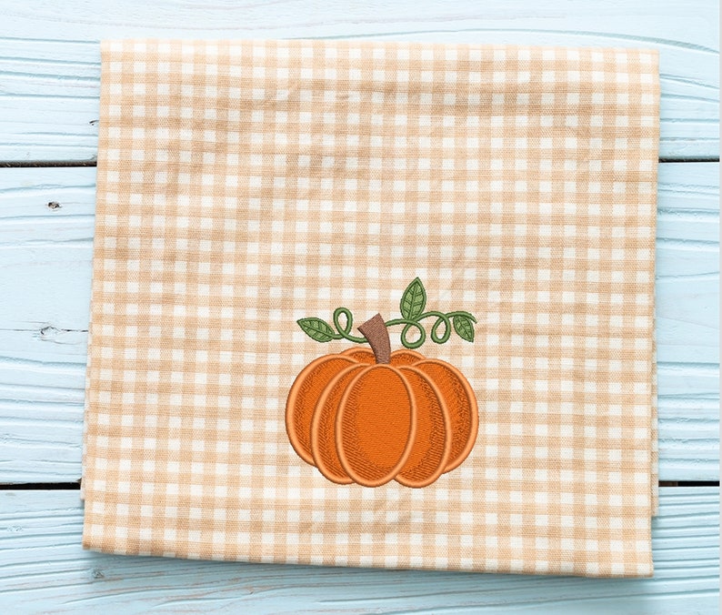 K&ouml;nnte beinhalten: Ein gestickter K&uuml;rbis in Braun und Orange mit einem gr&uuml;nen Stiel und Bl&auml;ttern auf einem beige-wei&szlig;en Gingham-Stoff.